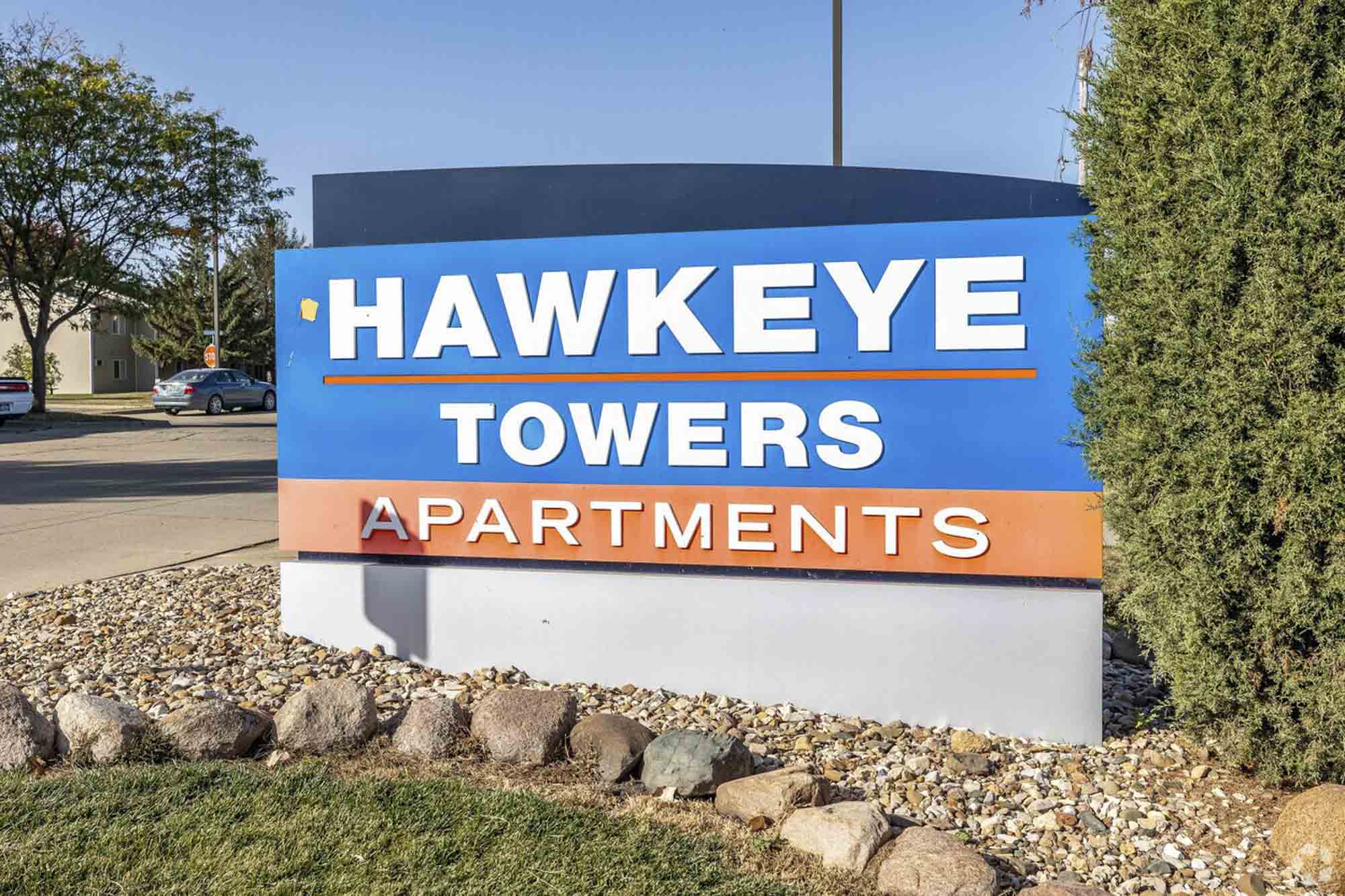 A large blue and orange sign for Hawkeye Towers Apartments set amidst rocks, grass, and a green bush, with residential buildings and cars visible in the background