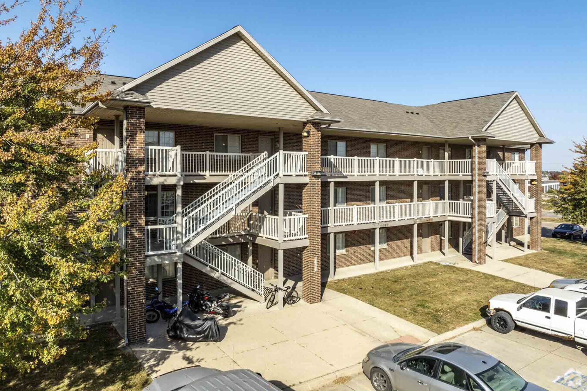 An angled view of a brick apartment building with external white staircases and balconies, surrounded by green grass and trees, with motorcycles and cars parked nearby