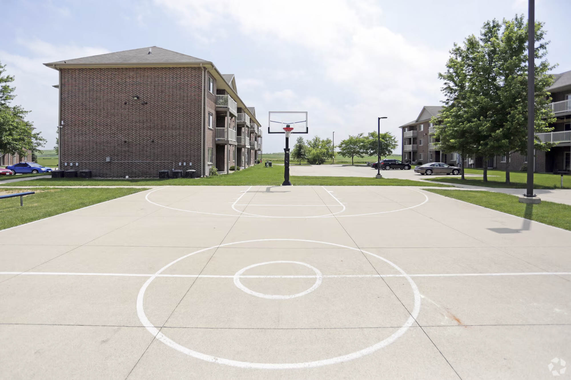 An outdoor basketball court with apartment buildings surrounding it and cars parked in the distance under a partly cloudy sky