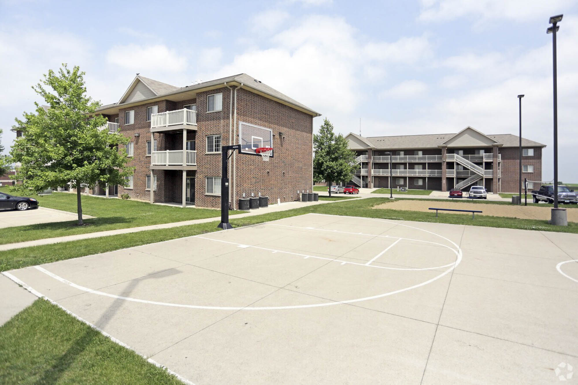 An outdoor basketball court with a hoop in the foreground, and multi-story brick apartment buildings with balconies and trees in the background under a blue sky